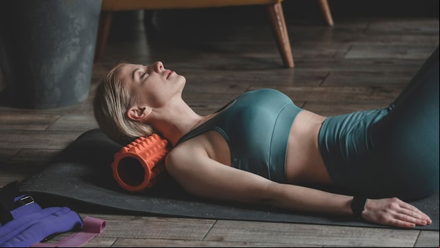 Mujer relajándose sobre una esterilla y un rodillo de masaje tras una jornada de entrenamientos. Está sobre una esterilla con ropa deportiva en plano medio.