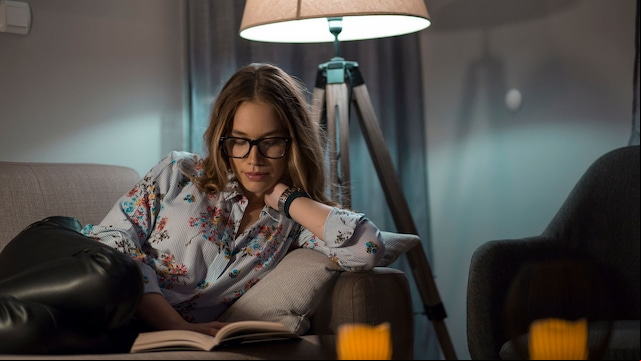 Mujer con gafas leyendo una novela en el sofá de su salón con la luz de una lámpara de pie detrás.