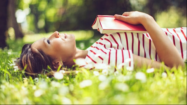 Mujer tumbada boca arriba sobre el césped, sonriendo, con un libro rojo entre las manos apoyado sobre el pecho